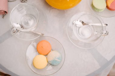 Bir kafede beyaz bir masa, üzerinde cam tabaklar ve kaşık dolu bardaklar var. Bir tabakta üç badem kurabiyesi var..