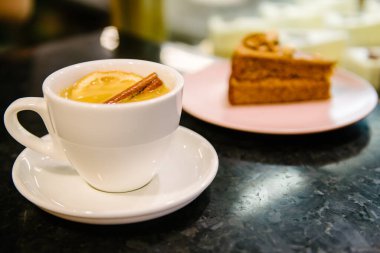 Beyaz bir tabakta beyaz bir fincanda tarçınlı meyve çayı. Tatlının arka planında pembe bir tabakta