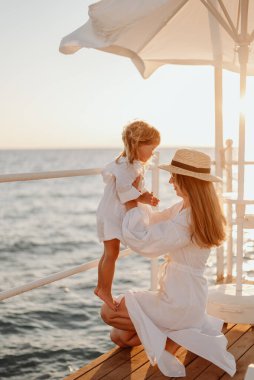 Genç bir kız, anne küçük kızını kollarına alıyor. Gece okyanusunun arka planında beyaz elbiseli kızlar. Hasır şapkalı kız.