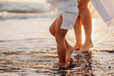 Yetişkin bir kadın ve bir kızın çıplak ayakları, beyaz elbiseli bir çocuk gün batımında okyanusta suyun üzerinde duruyor. Su damlaları güneşte parıldıyor