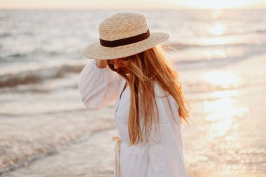 Uzun saçlı, hasır şapkalı ve beyaz elbiseli genç, güzel bir kız gün batımında deniz kıyısında olmaz. Bulanık arkaplan.