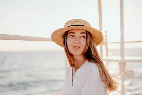 Stock image A beautiful young girl with long hair in a white dress and a straw hat smiles beautifully against the backdrop of the evening ocean.