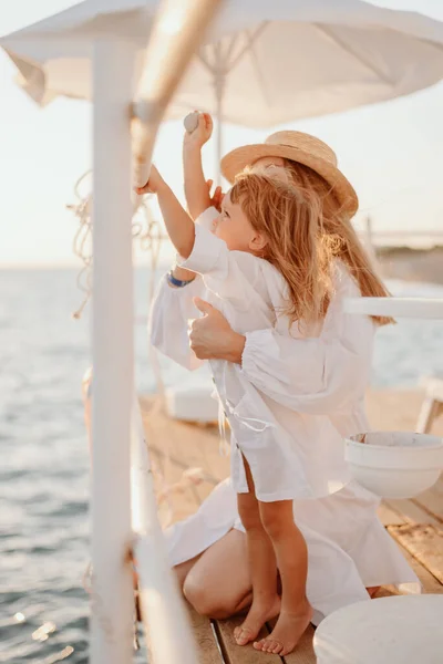 stock image A little girl, together with a young mother in white dresses and long hair, stand on the pier against the backdrop of the ocean at sunset