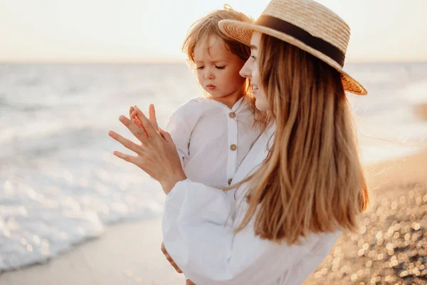 母親の腕の中に白いドレスを着た少女の小さな子供が 日没時の海を背景に母親の手に触れる ぼやけた背景 — ストック写真