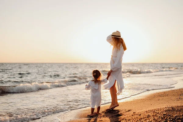 stock image A young girl, mother, with a little daughter in white dresses, walk on the water on the seashore at sunset.