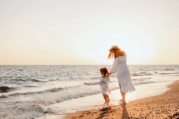 stock image A young mother holds her little daughter by the hand and together they walk along the ocean towards the sunset. Girls in white dresses and with long hair that develops the wind