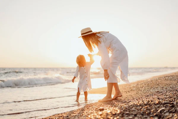 stock image A young mother holds her little daughter by the hand and together they walk along the ocean towards the sunset. Girls in white dresses and with long hair that develops the wind