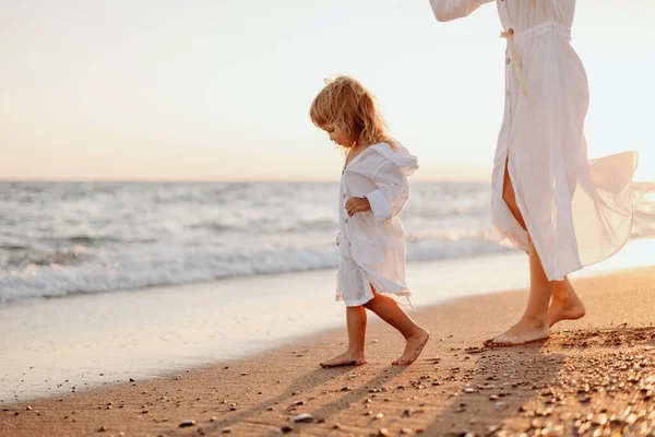 stock image A little girl and a young mother in white dresses and a hat are walking on the water on the ocean at sunset. Deserted beach.