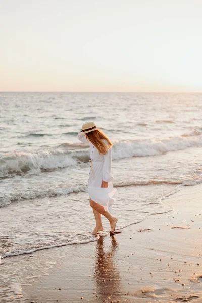 stock image A young, beautiful girl with long hair in a straw hat and a white dress dances on the water near the sea at sunset. Blurred background.