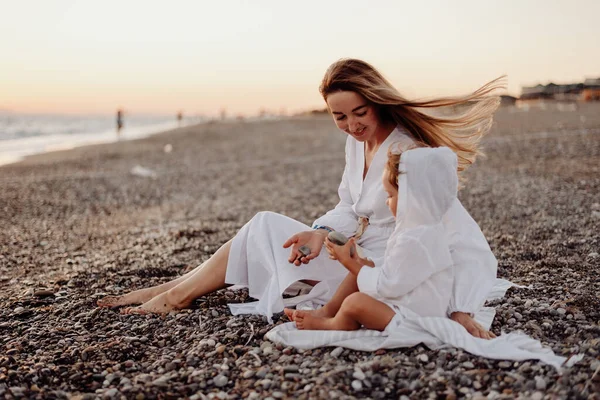 白いドレスを着た若い母親と娘が岩の多い海岸に座り 小石を手に持っています 海とピンクの夕日を背景に — ストック写真