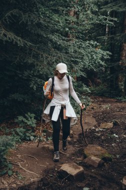 Sırt çantalı, beyaz şapkalı ve elinde sopayla gezen bir kız yeşil kozalaklı ormanların arasında dağ yolunda yürüyor.
