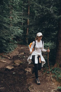 Sırt çantalı, beyaz şapkalı ve elinde sopayla gezen bir kız yeşil kozalaklı ormanların arasında dağ yolunda yürüyor.