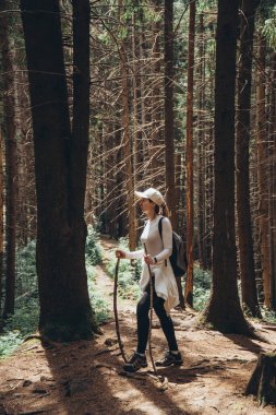 Uzun kozalaklı ağaçların arasında elinde sırıkla kayalık bir dağ yolunda duran bir gezgin..