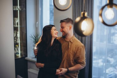 Evdeki evli bir çift sarılıyor ve birbirlerine gülümserken bakıyor. Kız, sırtı erkeğe dönük durur..