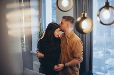 Bir adam ve bir kadın bir odada duruyor ve sarılıyorlar. Adam kızın elini tutuyor ve onu alnından öpüyor. Bulanık mutfak arka planı, ön plandaki röfleler