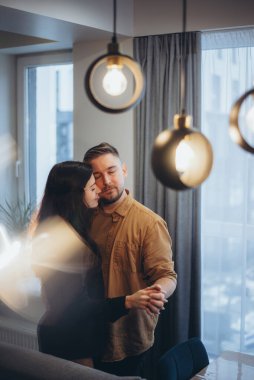 Evli bir çift evde dans ediyor. Bir kız ve bir erkek birbirlerine sarılıp yüzlerini bastırıyorlar. Bir erkek bir kızın elini tutar. Önplanda bulanık arkaplan ve renklendirmeler.