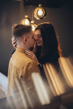 Bir kız ve bir erkek karşı karşıya duruyorlar. Adam nazikçe kızın belini tuttu. Kız, adamın yüzüne elleriyle dokunuyor. Arka plan bulanık. Ön planda parlama