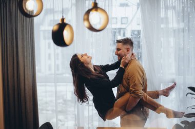 Pencerenin yanındaki bir otelde genç bir çift. Adam kızı kollarında tutuyor. Kız ellerini adamın boynunda tutuyordu. Gülümsüyorlar. Arka planda şehir bulanık. Ön planda parlama