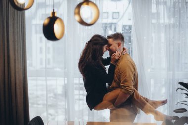 Pencerenin yanındaki bir otelde genç bir çift. Adam kızı kollarında tutuyor. Kız ellerini adamın boynunda tutuyordu. Gülümsüyorlar. Arka planda şehir bulanık. Ön planda parlama