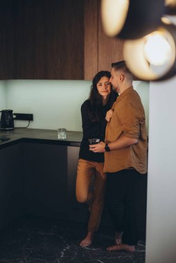 Mutfaktaki genç çift. Kız adamı gömleğinden tutuyor. Adam elinde bir bardak su tutuyor ve kıza bakıyor..