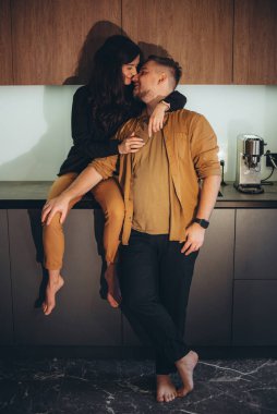 Mutfaktaki genç çift. Kız masanın üstüne oturur ve adama sarılmaya başlar. Bir adam çıplak ayakla yerde duruyor, bir kızı dizinden tutuyor ve gülümsüyor.