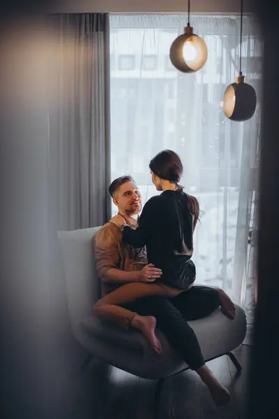 The girl sits on the park in an intimate cowgirl position on a chair. A young, married couple in love. The guy and the girl look tenderly at each other. There is a panoramic window in the background. Blurred foreground.