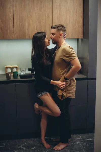 A young couple is standing in the kitchen. A girl in shorts with bare legs. The girl takes off the guy\'s shirt and smiles tenderly. On the background there is a tabletop and two coffee cups