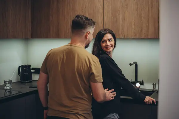 A young couple is standing in the kitchen. The guy sneaks up behind the girl and touches her waist with his hand. The girl turned to the guy and smiled