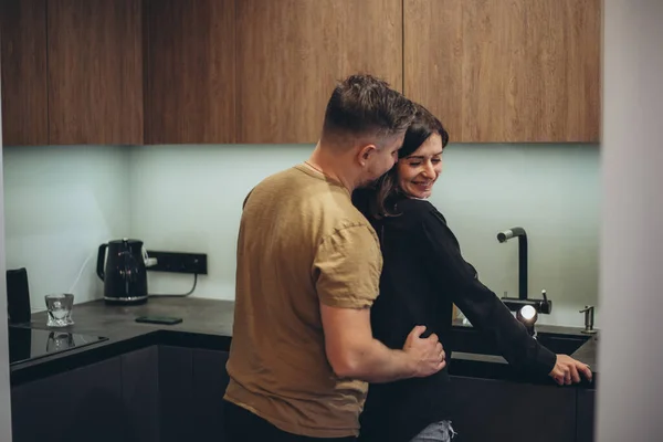 A young couple is standing in the kitchen. The guy sneaks up behind the girl and touches her waist with his hand. The girl turned to the guy and lowered her eyes in embarrassment