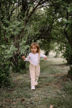 Küçük şık bir kız, bej pantolonlu ve beyaz bluzlu bir çocuk, uzun yeşil çalılıklardan bir tünelde koşuyor.