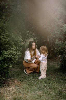Küçük şık bir kız, bir çocuk ve annesi çalıların arasındaki yeşil bir çimenlikte oturdu. Anne kızına sarılıyor. Kızın elinde bir sepet ve çiçek açan bir elma ağacı dalı var..