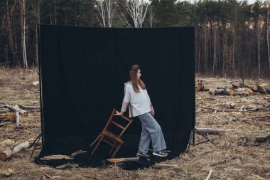 Güzel üzgün bir kız, kara bir arka planda, ormansızlaşmanın ortasında yerde duruyor. Her yerde ağaç kütükleri var. Ormansız bir orman. Doğayı yok etti. Üzgün kız.