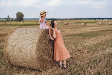 Anne ve küçük kızı mavi gökyüzüne karşı hasat edilmiş buğday tarlasında. Kız bir saman balyasının üzerinde oturuyor. Annem bir saman balyasına yaslandı ve gözlerini kapattı..