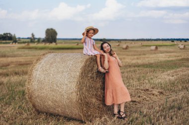 Anne ve küçük kızı hasat edilen buğday tarlasında saman balyalarının arka planında. Kız bir saman balyasının üzerinde oturuyor. Anne kızının yanında, kızının elini tutuyor.