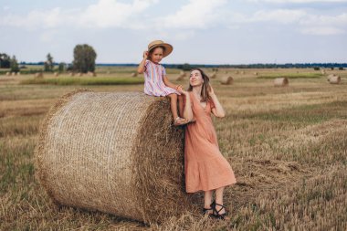 Anne ve küçük kızı hasat edilen buğday tarlasında saman balyalarının arka planında. Kız bir saman balyasının üzerinde oturuyor. Annem kızına bakıyor..