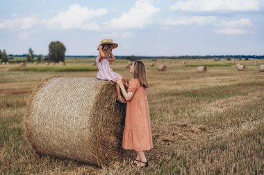 Hasat edilen buğday tarlası. Küçük bir kız bir saman balyasına oturur ve şapkasını kafasına takar. Mercan elbiseli yetişkin bir anne yakınlarda duruyor ve kızına bakıyor.
