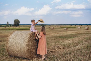 Hasat edilen buğday tarlası. Küçük bir kız bir saman balyasının üzerinde oturuyor. Mercan elbiseli yetişkin bir anne yakınlarda durup kızına bakar. Bir kız annesine şapka takmaya çalışıyor.