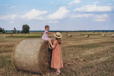 Hasat edilen buğday tarlası. Küçük bir kız bir saman balyasının üzerinde oturuyor. Mercan elbiseli yetişkin bir anne yakınlarda durup kızına bakar. Bir kız annesinin kafasına şapka takar.