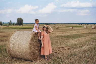 Hasat edilen buğday tarlası. Küçük bir kız bir saman balyasının üzerinde oturuyor. Mercan elbiseli yetişkin bir anne yakınlarda durup kızına bakar. Kız annesinin kafasına bir şapka takıyor. Annem gülümser.