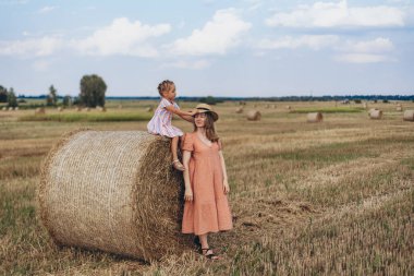 Hasat edilen buğday tarlası. Küçük bir kız bir saman balyasının üzerinde oturuyor. Mercan elbiseli yetişkin bir anne yakınlarda durup kızına bakar. Bir kız annesinin kafasına şapka takar.