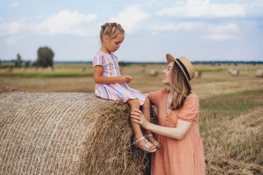 Hasır şapkalı genç bir anne ve hasat edilen buğday tarlasında iletişim kuran genç bir kız, saman balyasında oturan bir kız.