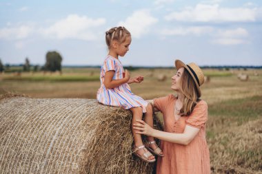 Hasır şapkalı genç bir anne ve hasat edilen buğday tarlasında iletişim kuran genç bir kız, saman balyasında oturan bir kız.