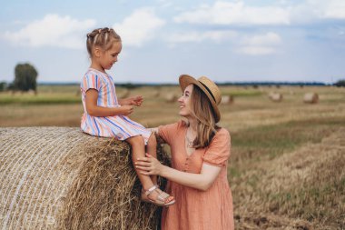 Hasır şapkalı genç bir anne ve hasat edilen buğday tarlasında iletişim kuran genç bir kız, saman balyasında oturan bir kız.