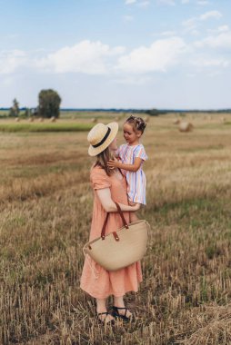 Uzun elbiseli ve hasır şapkalı genç bir anne küçük kızını elinden tutar. Her ikisi de hasat edilen buğday tarlasında yürür. Bir saman balyasının arka planında