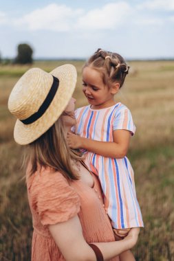 Hasır şapkalı ve hasır çantalı genç bir anne kucağında küçük bir kızı tutuyor. Her ikisi de hasat edilen buğday tarlasında duruyor. Bir saman balyasının arka planında