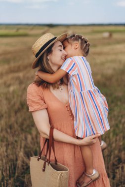 Hasır şapkalı ve hasır çantalı genç bir anne kucağında küçük bir kızı tutuyor. Her ikisi de hasat edilen buğday tarlasında duruyor. Bir saman balyasının arka planında