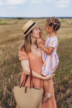 Hasır şapkalı ve hasır çantalı genç bir anne kucağında küçük bir kızı tutuyor. Her ikisi de hasat edilen buğday tarlasında duruyor. Bir saman balyasının arka planında