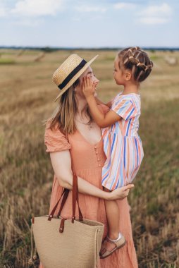 Hasır şapkalı ve hasır çantalı genç bir anne kucağında küçük bir kızı tutuyor. Her ikisi de hasat edilen buğday tarlasında duruyor. Bir saman balyasının arka planında