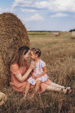 Anne ve kızı hasat edilen buğday tarlasında saman balyasının yanında oturuyor. Diğer balyaların arka planına karşı. Anne kızıyla yüzleşmek için arkasına yaslanıyor.