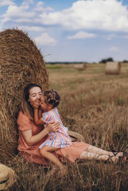 Anne ve kızı hasat edilen buğday tarlasında saman balyasının yanında oturuyor. Diğer balyaların arka planına karşı. Kızın, annenin yüzüne bastırılmış.